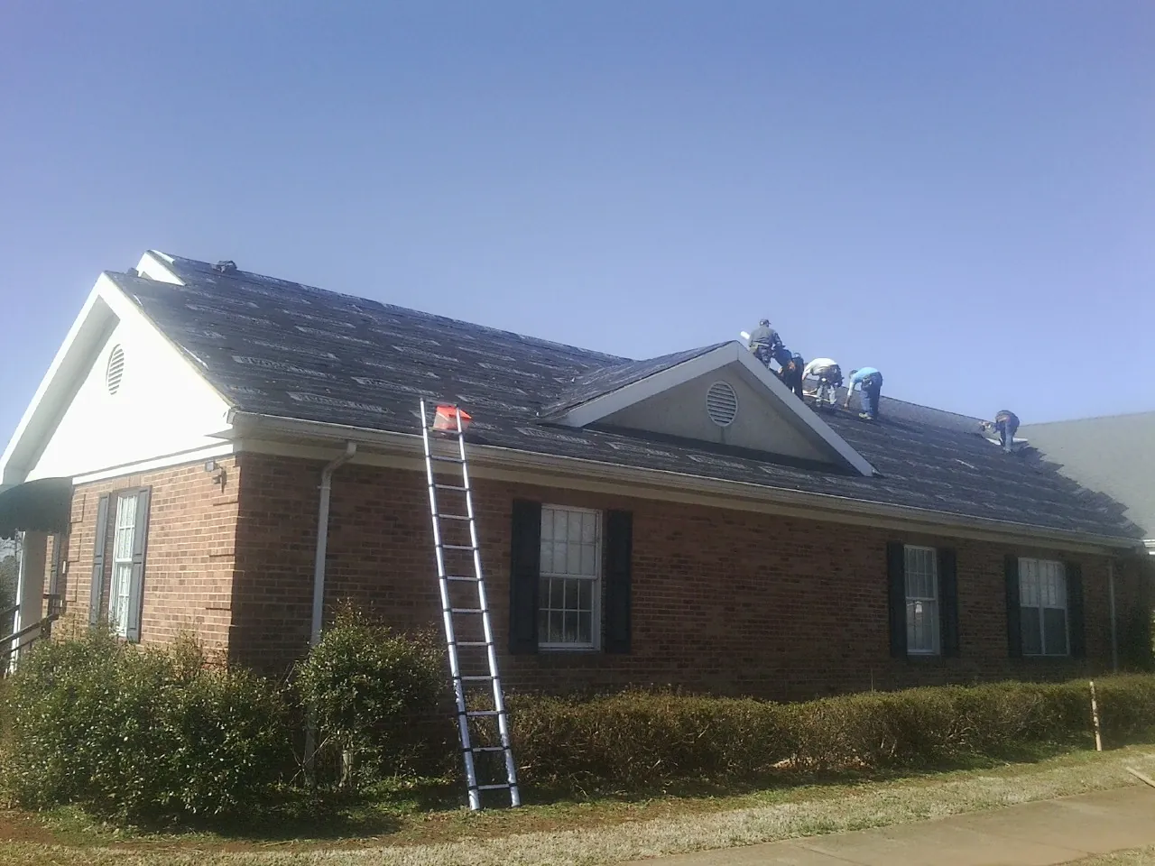 Crew tearing shingles off a brick ranch roof with a ladder set — Emergency Roof Repair work by our crew in Albin