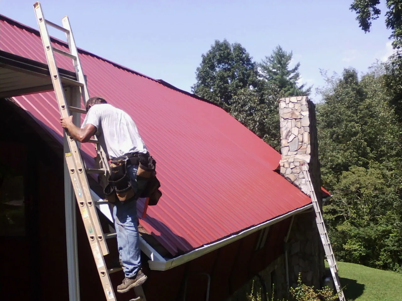 Roofer on a ladder beside a red metal roof and stone chimney — Soffit & Fascia Repair by Roofing Brothers in Albin